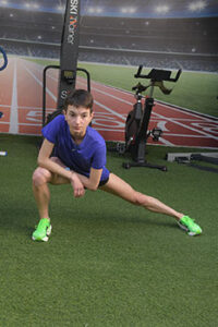 Νεαρή αθλήτρια εκτελεί άσκηση διάτασης με πλευρικό άνοιγμα ποδιών σε εσωτερικό αθλητικό χώρο με φόντο ψηφιακό στάδιο. Φοράει μπλε αθλητική στολή και πράσινα αθλητικά παπούτσια Asics. Στο βάθος διακρίνεται προσομοιωτής σκι Ski Trainer και ποδήλατο γυμναστικής. Η άσκηση χρησιμοποιείται για βελτίωση ευλυγισίας, κινητικότητας και προθέρμανση πριν την προπόνηση. 