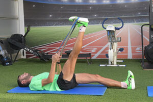 Αθλητής εκτελεί ισομετρική άσκηση οπίσθιων μηριαίων με εξοπλισμό Nordic Curl σε γυμναστήριο με φόντο ψηφιακό στάδιο. Ο αθλητής βρίσκεται σε ύπτια θέση σε γαλάζιο στρώμα άσκησης με τα πόδια ασφαλισμένα στη συσκευή, φορώντας πράσινα αθλητικά παπούτσια. Η άσκηση χρησιμοποιείται για ενδυνάμωση οπίσθιων μηριαίων, πρόληψη τραυματισμών και αξιολόγηση εκκεντρικής δύναμης. 