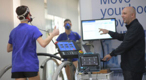 Νεαρή αθλήτρια με μάσκα αναπνοής υποβάλλεται σε δοκιμασία VO2 Max σε διάδρομο υπό την επίβλεψη ειδικού φυσιολόγου άσκησης. Ο επαγγελματίας εξηγεί τα αποτελέσματα που εμφανίζονται σε οθόνη υπολογιστή.